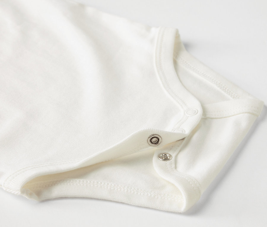 Detail bieleho detského body s krátkym rukávom a patentkami.