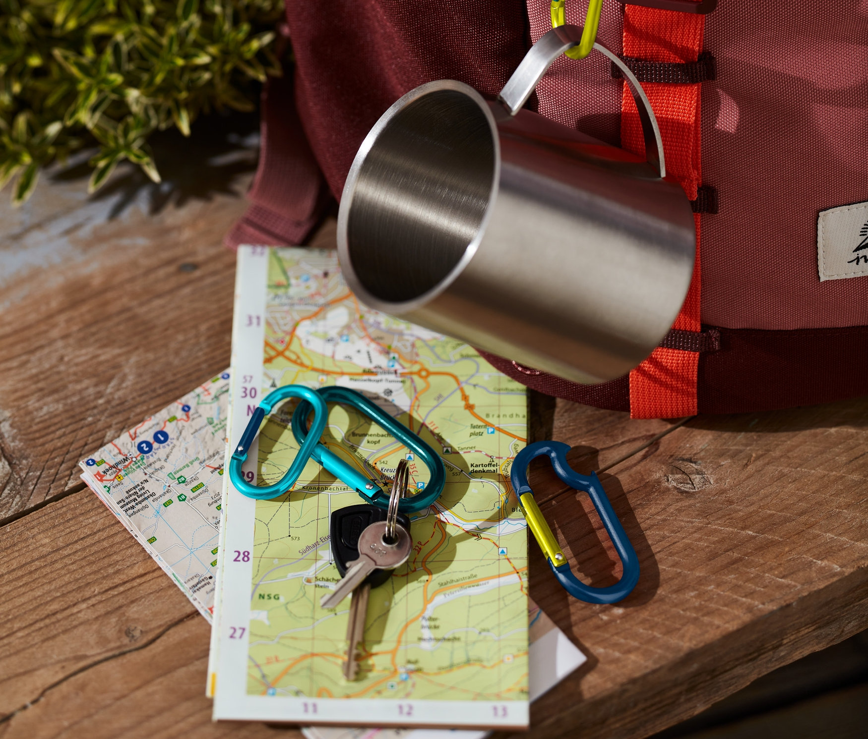 Nerezový hrnček visí na batohu pomocou jednej zo 4 karabín. Na mape ležia kľúče s ďalšou karabínou.