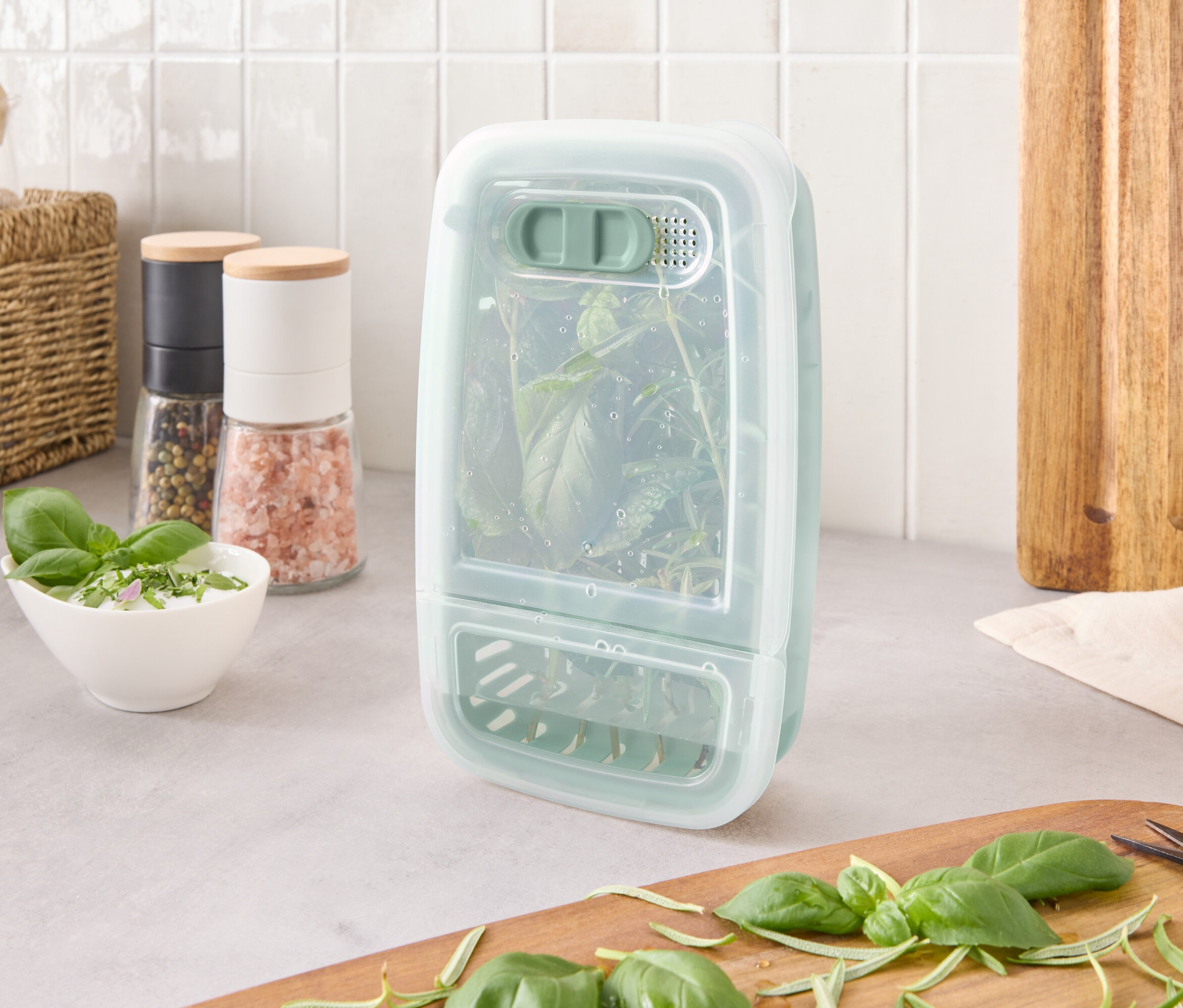 Nádoba na uchovávanie byliniek s bylinkami vnútri stojí na linke vedľa ingrediencií a dosky na krájanie.