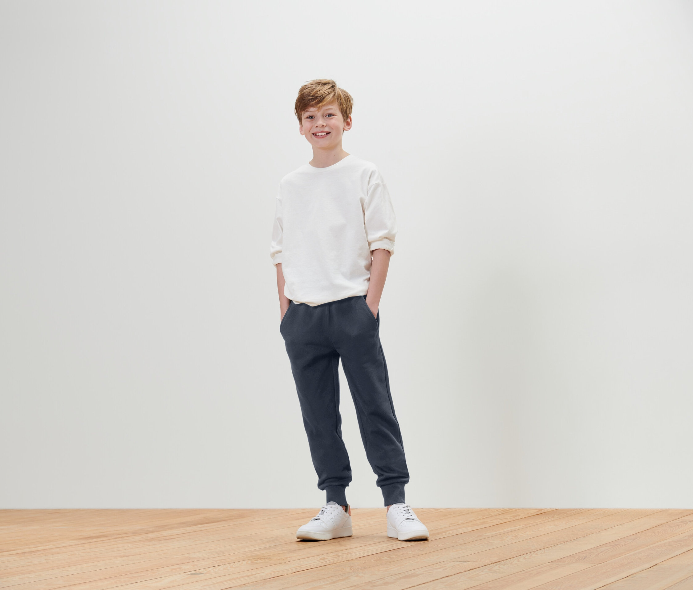 Chlapec stojí s rukami vo vreckách a má na sebe biele tričko, modré tepláky a biele tenisky.