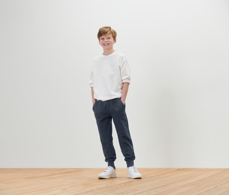 Chlapec stojí s rukami vo vreckách a má na sebe biele tričko, modré tepláky a biele tenisky.