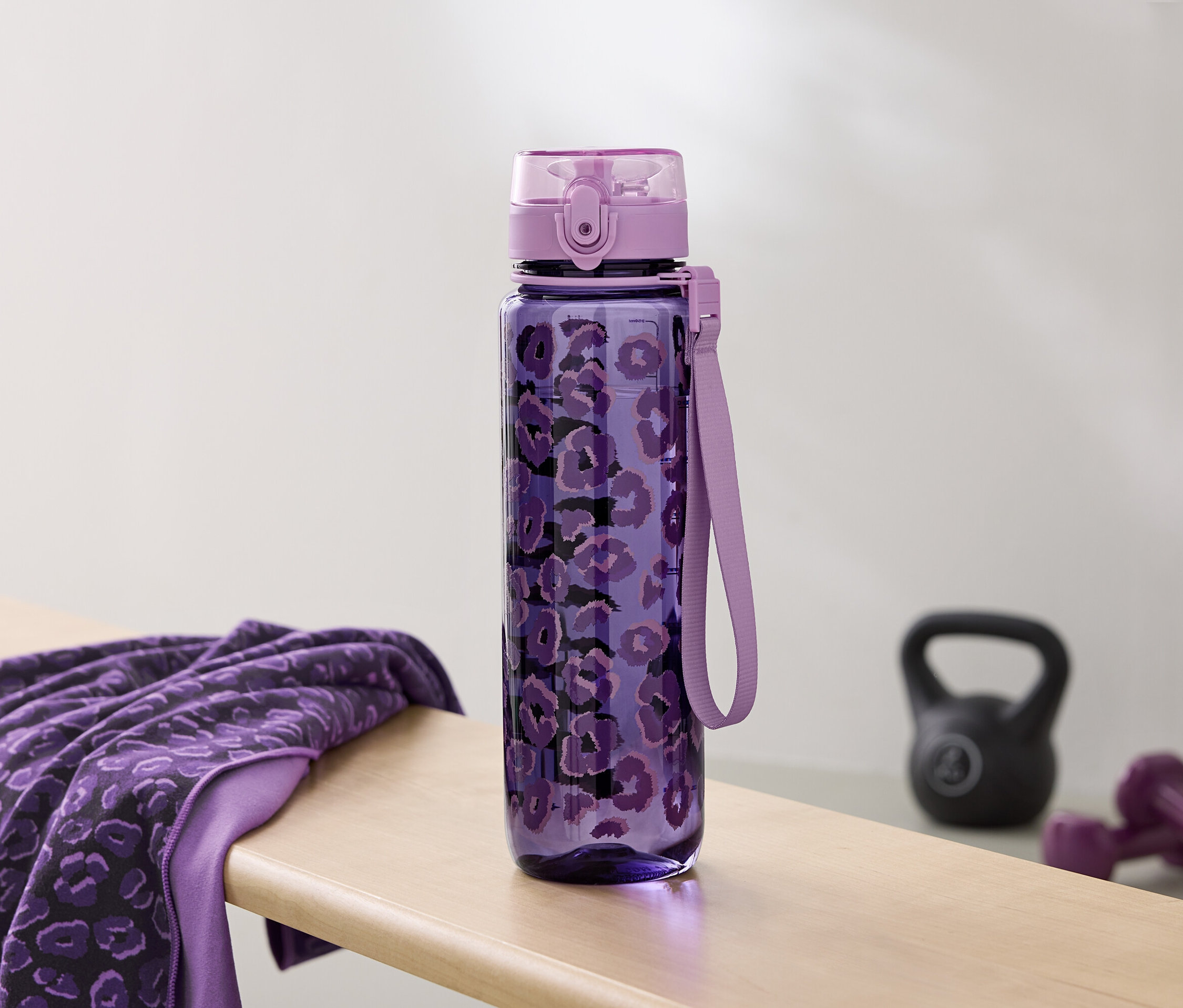 Fialová fľaša na vodu s potlačou leží vedľa uteráka a činiek na lavičke.