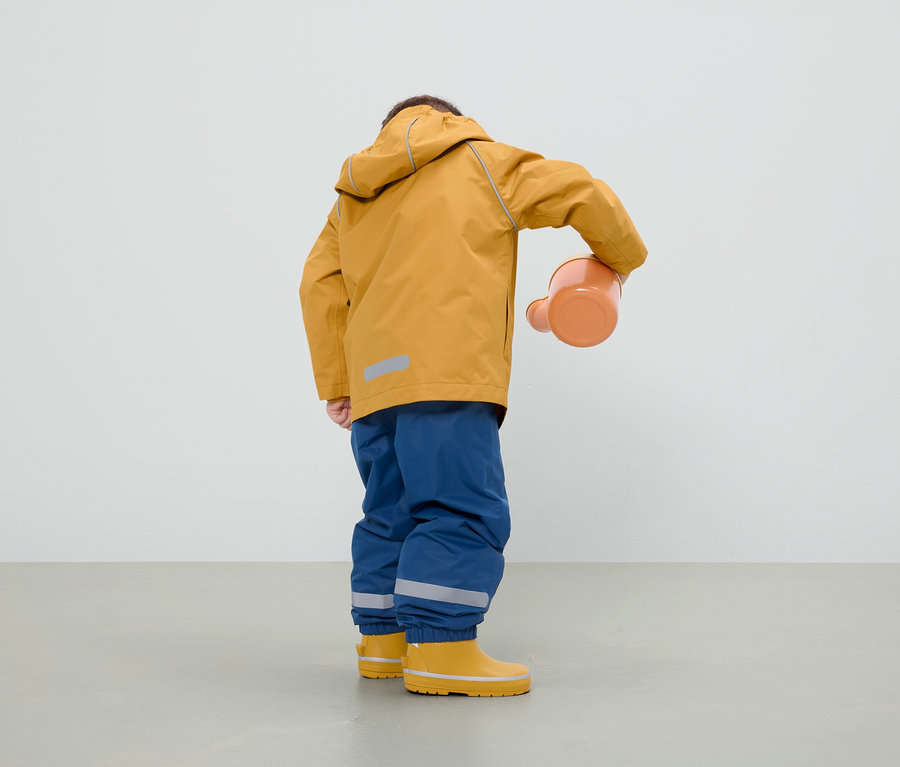 Chlapec v horčicovej bunde, modrých nohaviciach a žltých gumákoch drží oranžovú kanvicu.