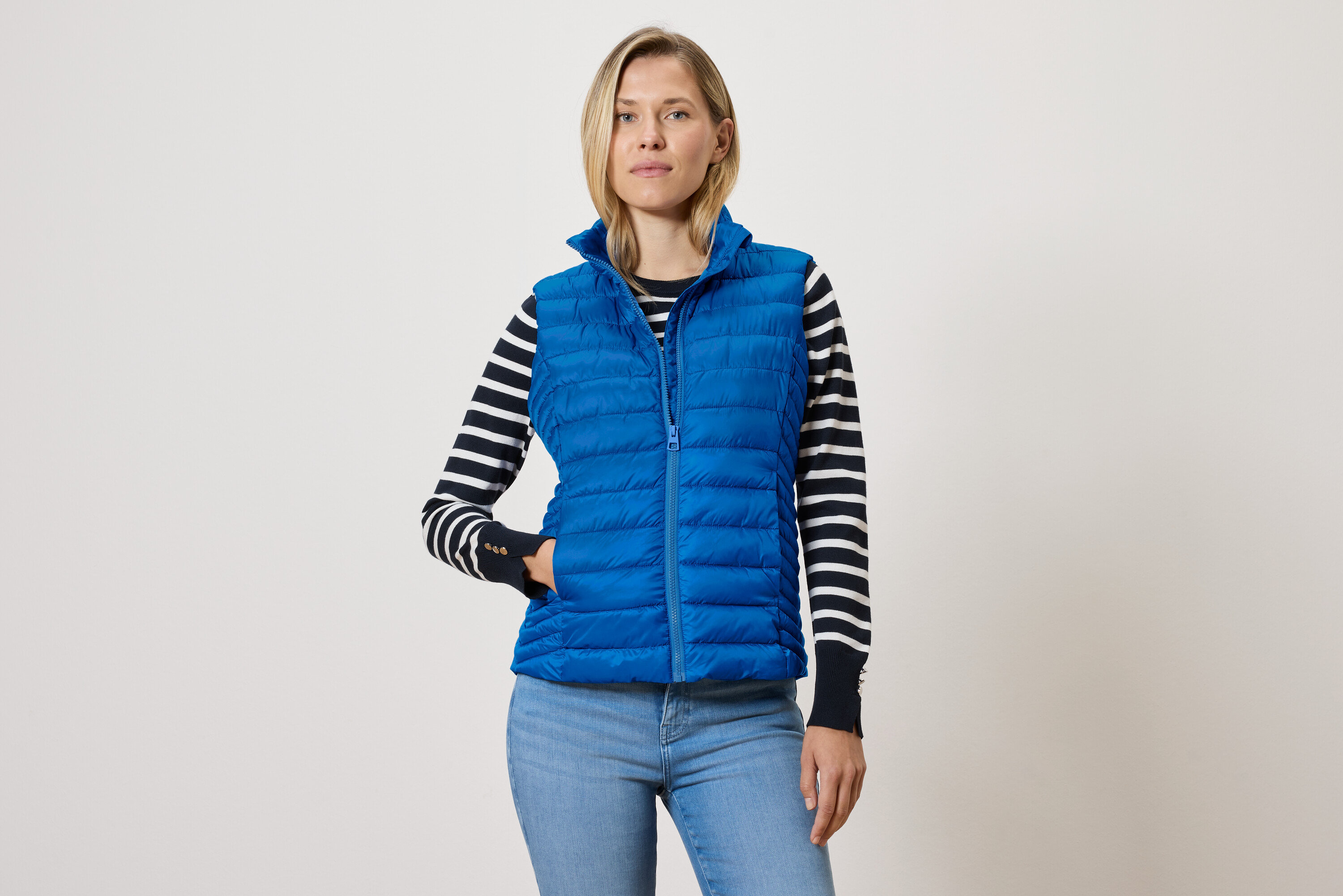 Žena v modrej prešívanej veste so zipsom, svetri s čiernymi a bielymi pruhmi a modrých džínsoch.
