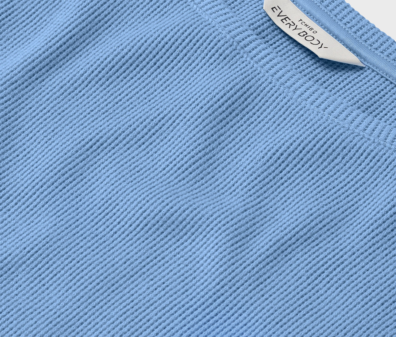 Detail svetlomodrej textílie s rebrovanou štruktúrou a bielym štítkom s nápisom EVERYBODY.