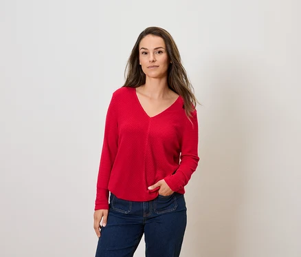 Žena s hnedými vlasmi pózuje v červenom svetri s dlhým rukávom a modrých džínsoch.