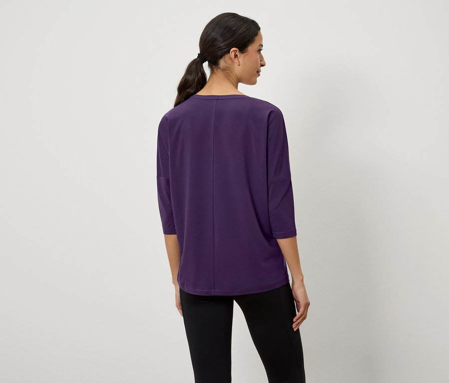 Žena s tmavými vlasmi stiahnutými do copu stojí chrbtom a má na sebe fialové tričko s trojštvrťovými rukávmi a čierne legíny.