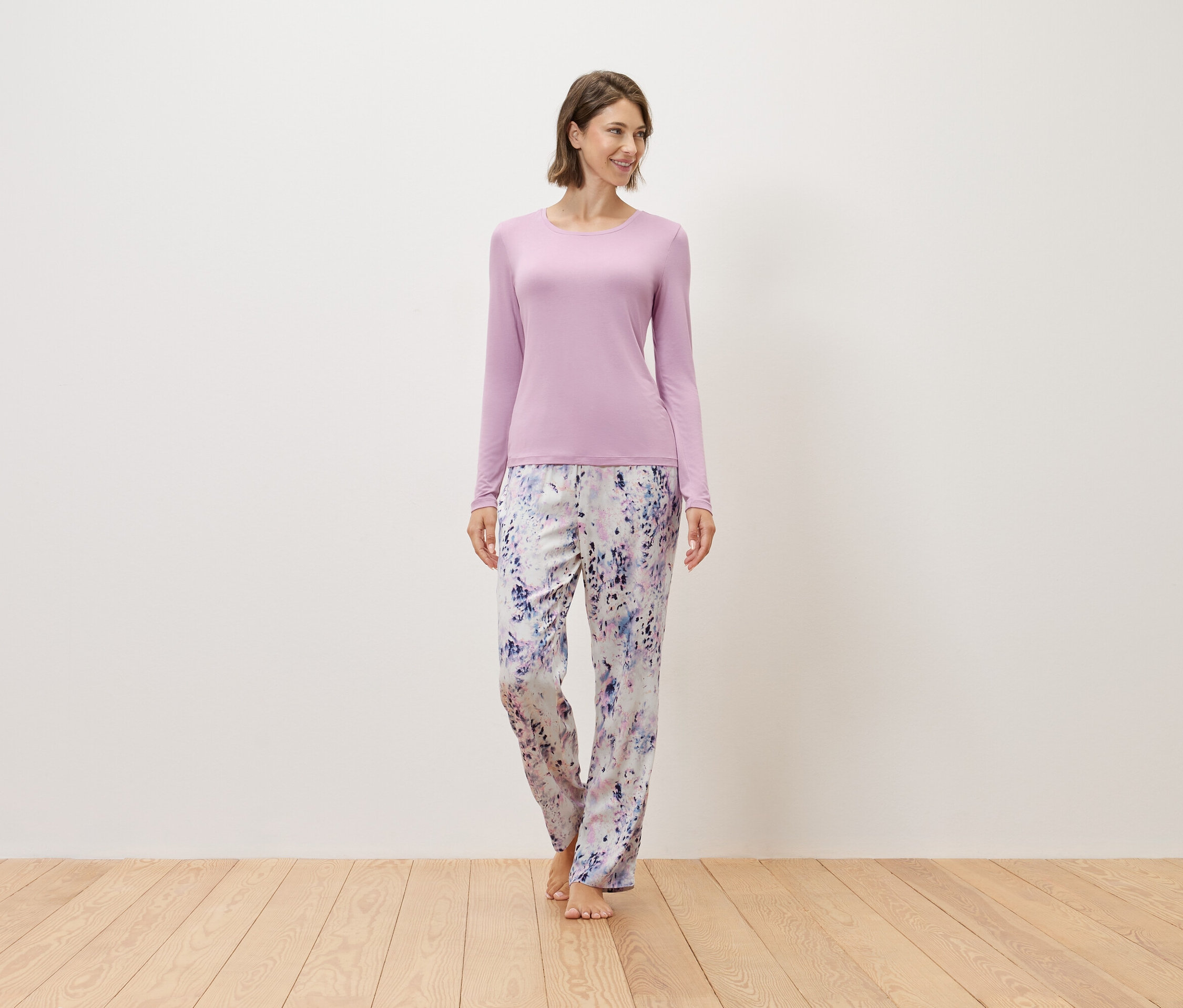 Žena stojí naboso na drevenej podlahe, má na sebe fialový sveter a vzorované nohavice.