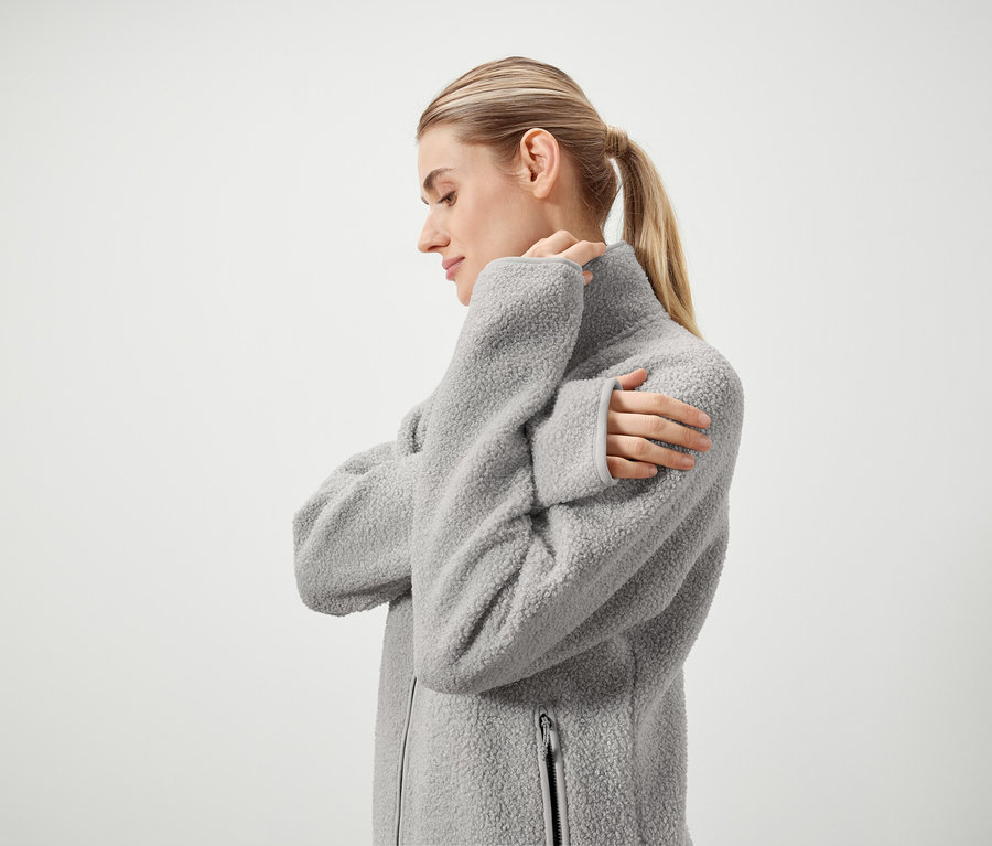 Profil ženy s blond vlasmi stiahnutými do copu, ktorá má na sebe sivú chlpatú bundu so zipsom a stojí pred bielym pozadím.