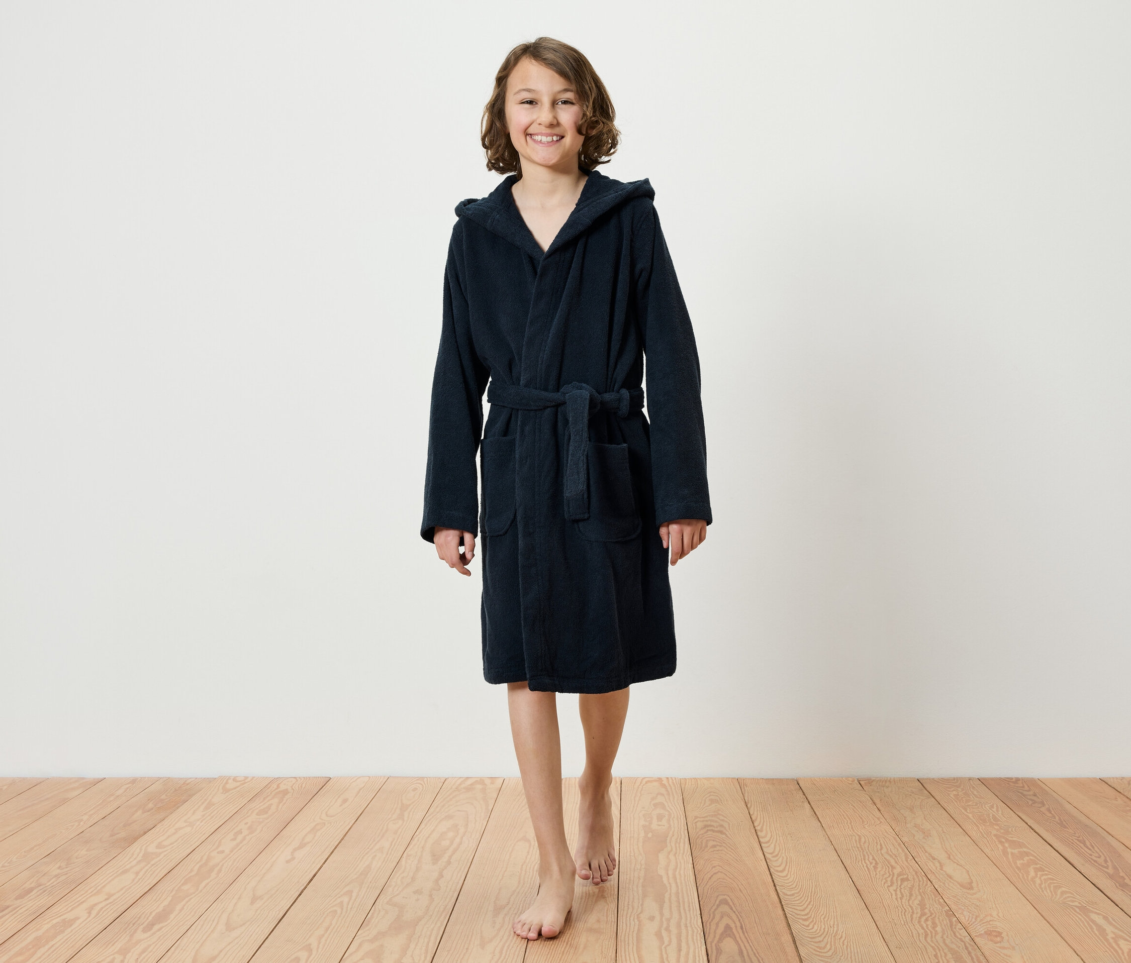 Usmievajúce sa dospievajúce dievča kráča naboso po drevenej podlahe a má na sebe tmavomodrý župan s kapucňou.