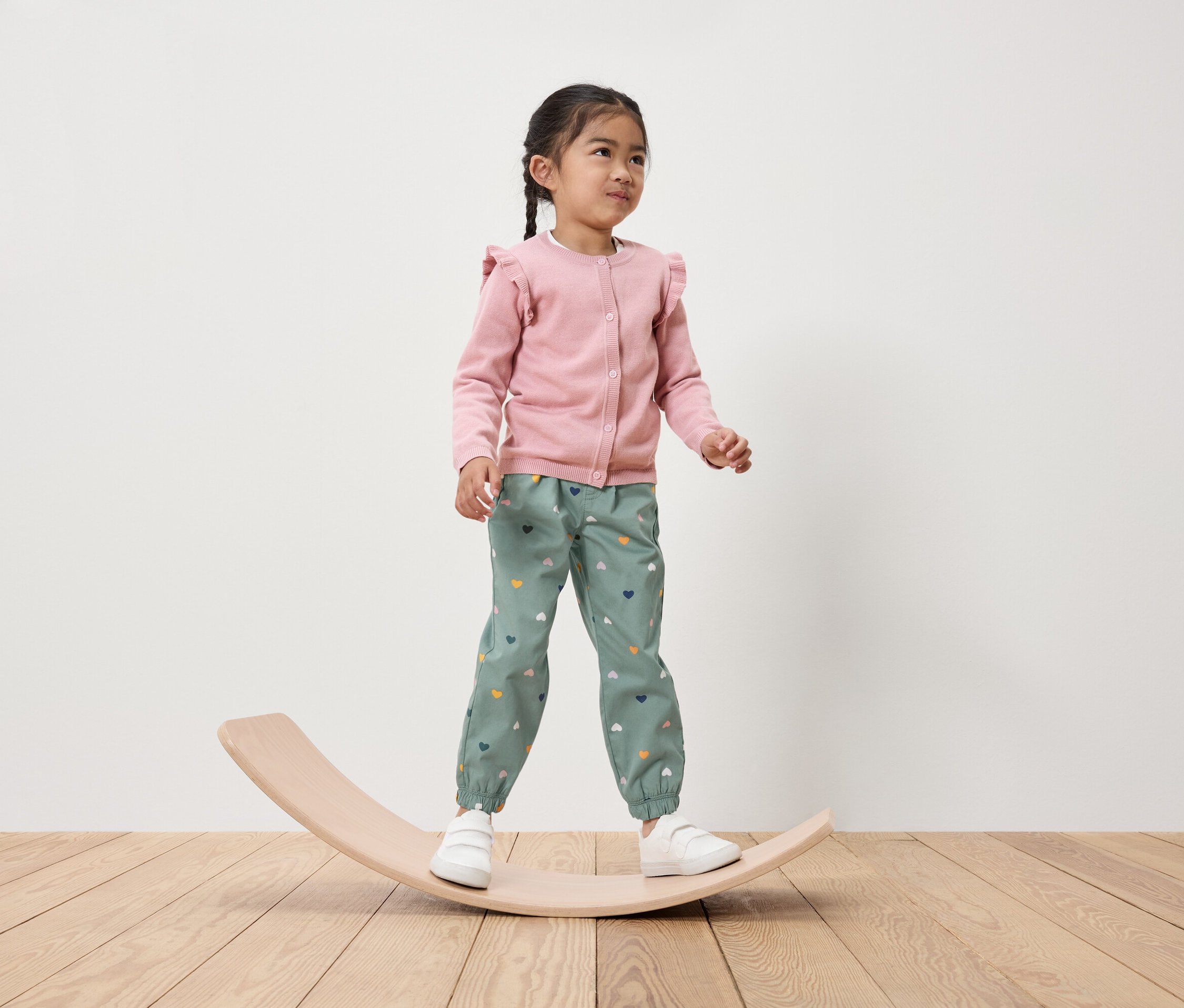 Dievčatko stojí na drevenej balančnej doske, má na sebe ružový kardigán a zelené nohavice so srdiečkami.