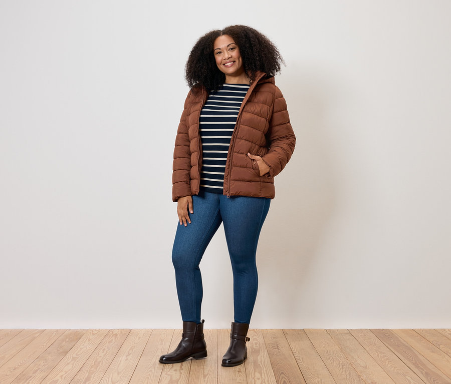 Žena s kučeravými vlasmi pózuje v hnedej prešívanej bunde, modrých džínsoch a hnedých topánkach.