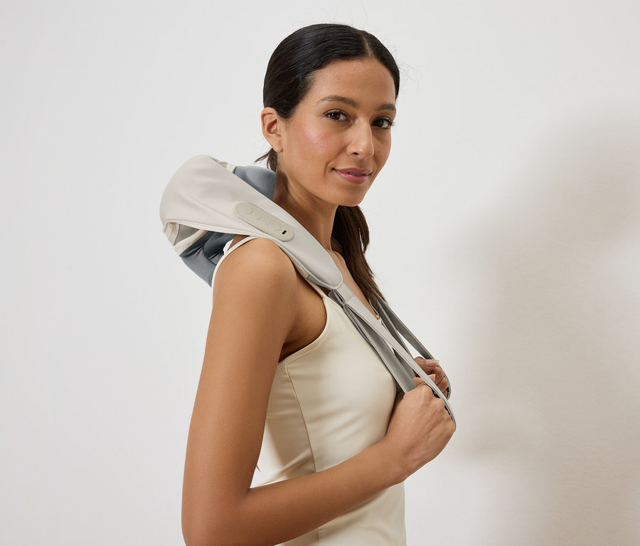 Žena s masážnym prístrojom na krk v sivej a béžovej farbe na pleciach, zariadenie drží v rukách.