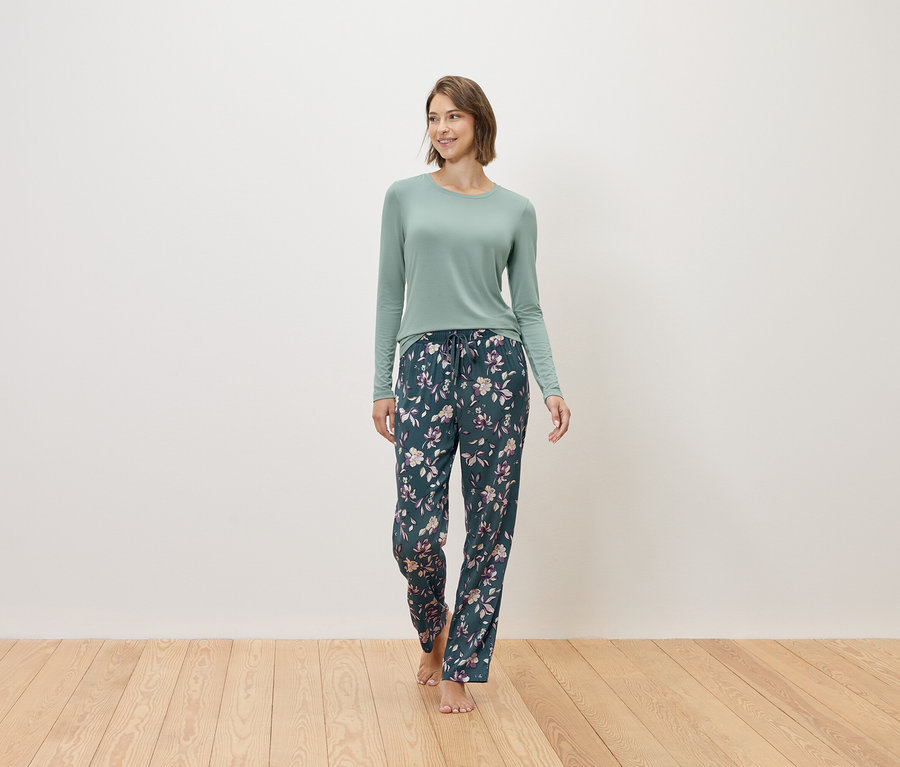 Žena kráča naboso a má na sebe mätový sveter a tmavomodré pyžamové nohavice s kvetinovým vzorom.