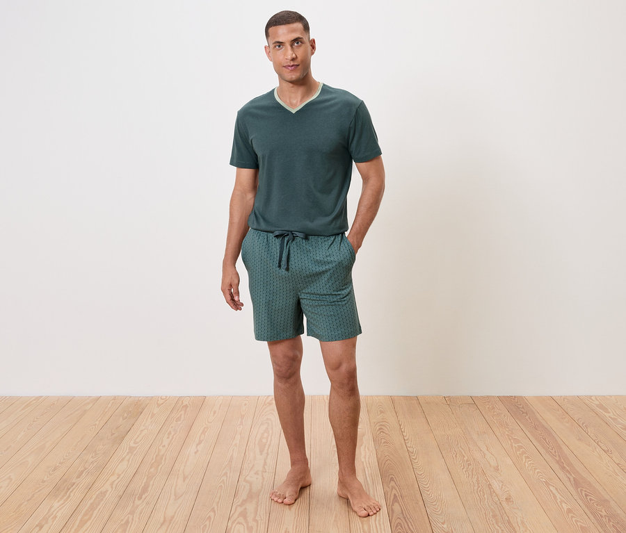 Muž stojí bosý na drevenej podlahe a má na sebe zelené pyžamo s krátkym rukávom a šortky.