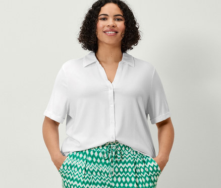 Žena stojí a má ruky vo vreckách trojštvrťových džersejových nohavíc s potlačou a bieleho blúzkového trička s gombíkovou légou.