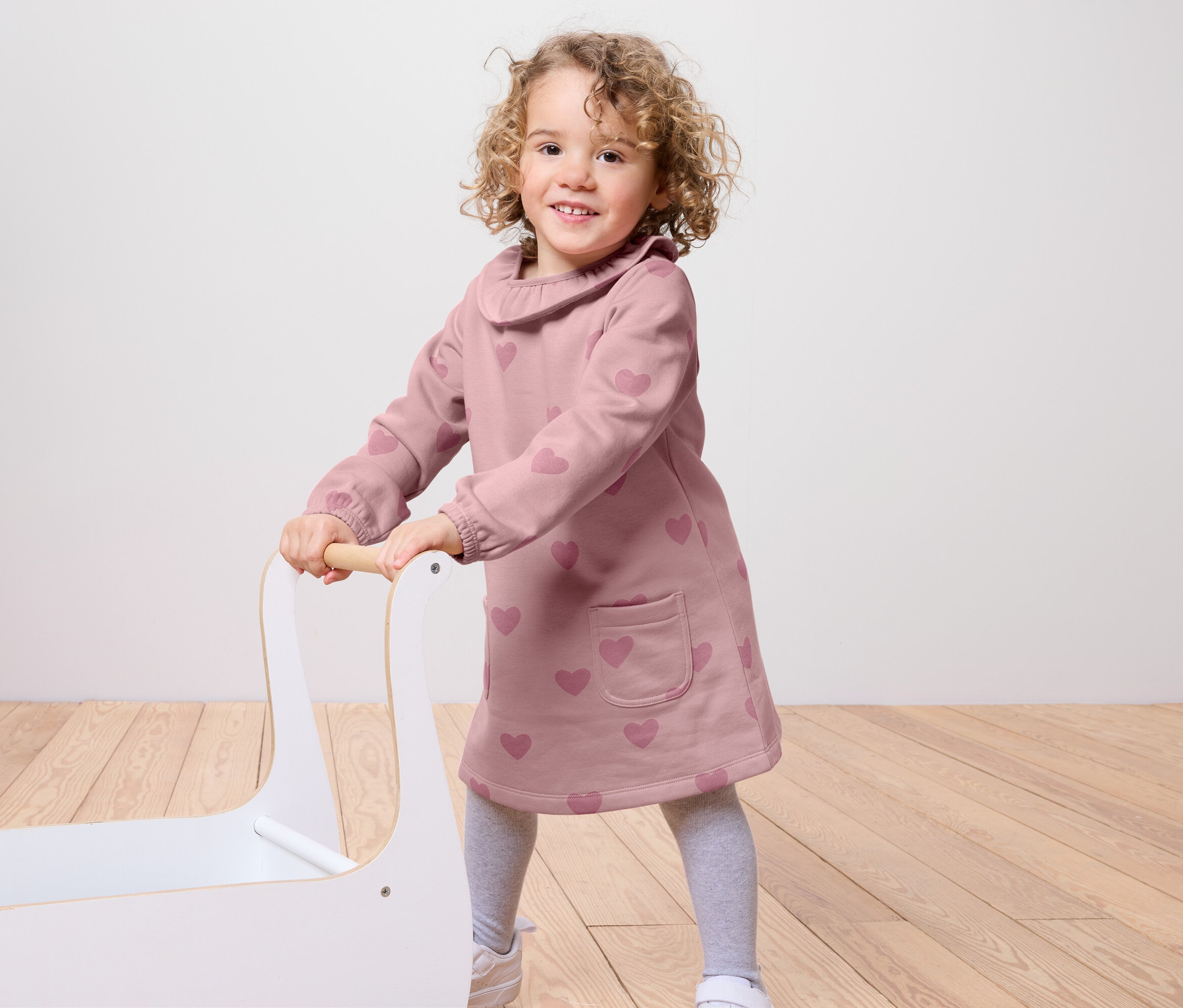 Usmievajúce sa dievčatko tlačí biely vozík na hračky. Má na sebe ružové šaty so srdiečkami, sivé legíny a biele tenisky.