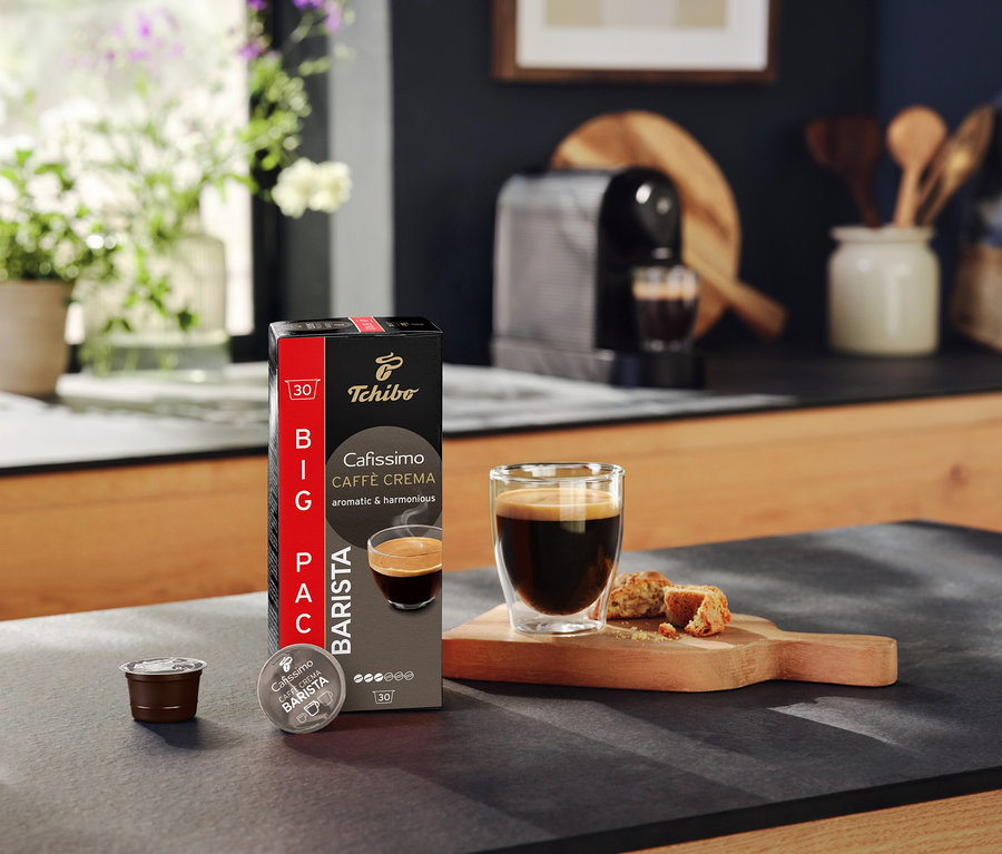 Na kuchynskej linke stojí balenie Tchibo Cafissimo Caffè Crema, kapsula, šálka kávy a pečivo.