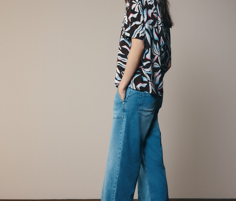 Žena v vzorovanej blúzke a rozšírených džínsoch, pozerajúca sa nabok.