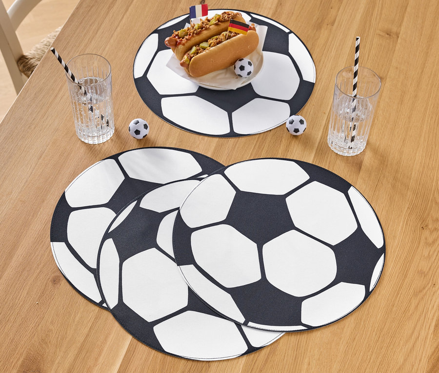 Dva hot dogy s vlajkami na tanieri s futbalovým vzorom, vedľa poháre so slamkami a futbalová dekorácia na drevenom stole.