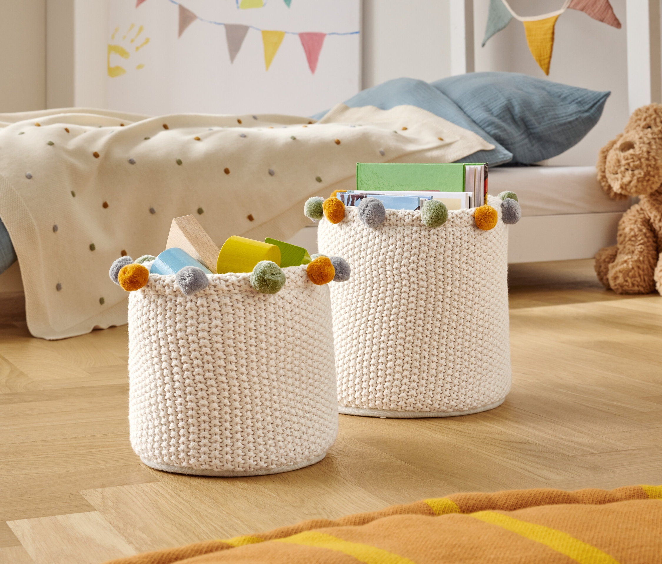 Dva pletené úložné koše s farebnými brmbolcami stoja na drevenej podlahe v detskej izbe.