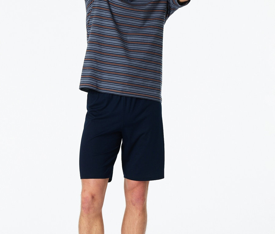 Muž stojí naboso so zdvihnutými rukami v pruhovanom tričku a modrých šortkách na bielom pozadí.