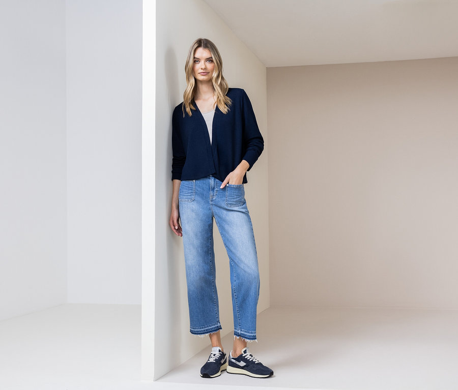 Žena sa opiera o stenu, má na sebe tmavomodrý kardigán, modré džínsy a tmavomodré topánky.