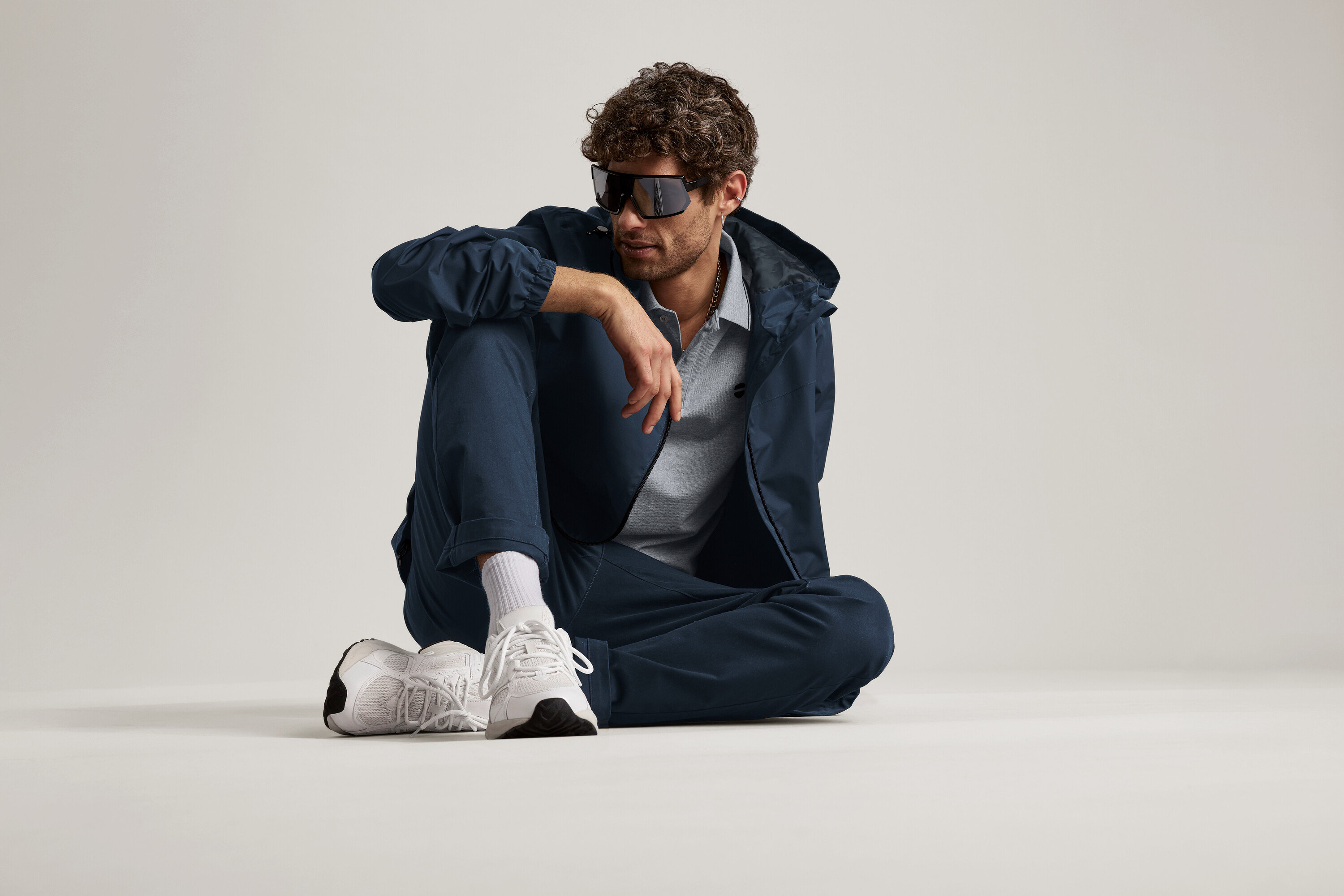 Muž sedí na zemi v námorníckych chino nohaviciach, bunde a slnečných okuliaroch.