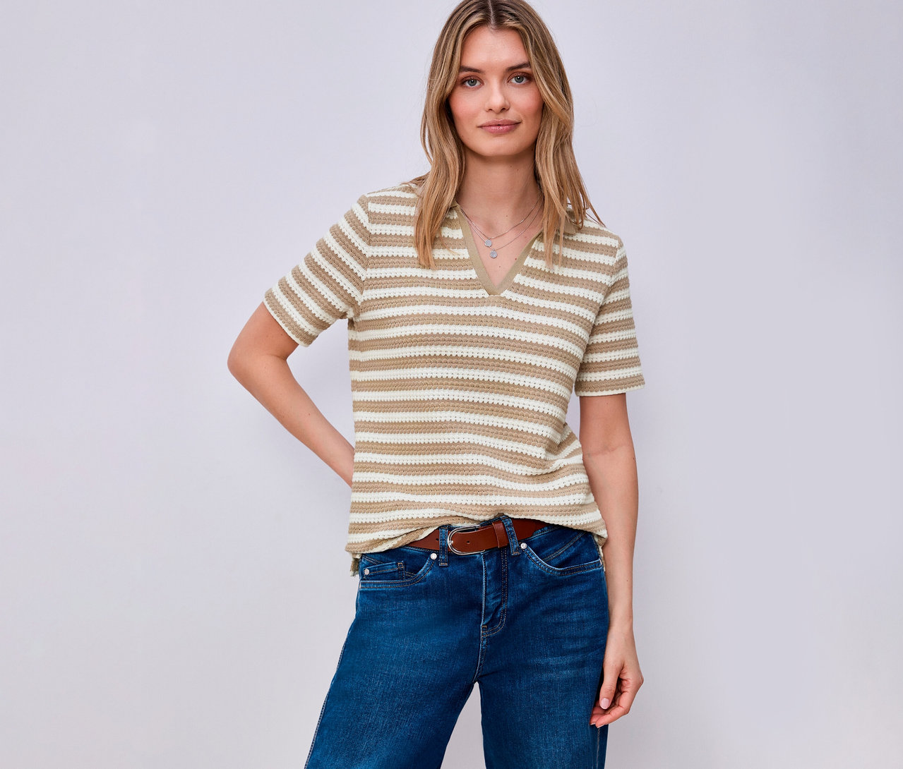 Žena s hnedým opaskom a v džínsoch má na sebe prúžkované tričko z úpletu Street One. Pózuje na svetlom pozadí.