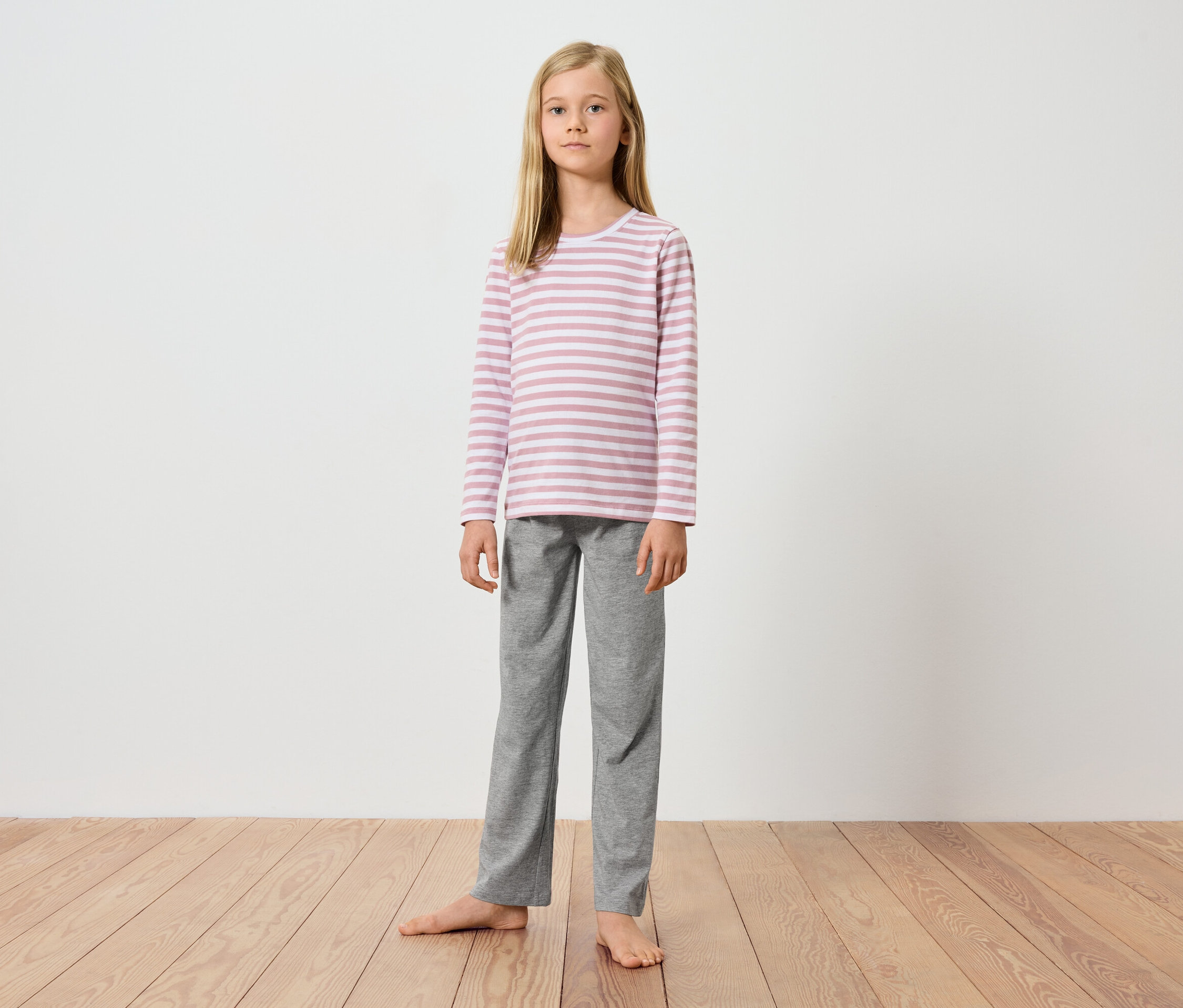 Mladé dievča stojí bosé na drevenej podlahe pred bielou stenou. Má na sebe ružovo-biele pruhované tričko s dlhým rukávom a sivé nohavice.