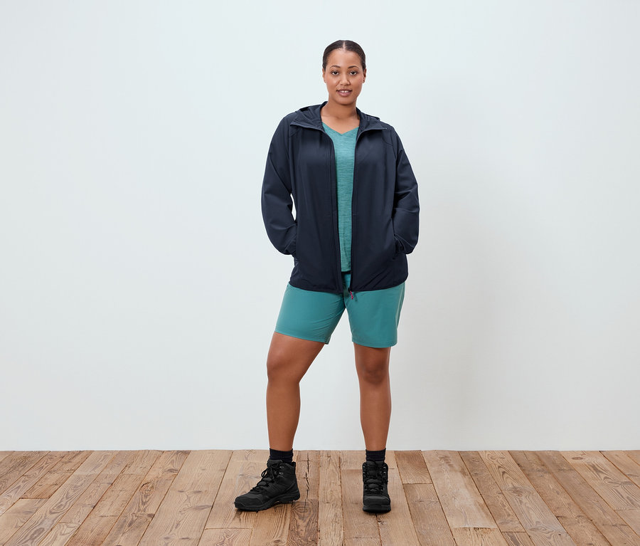 Žena stojí s rukami vo vreckách pred bielou stenou. Má na sebe tmavomodrú bundu, tyrkysové tričko, tyrkysové šortky a čierne topánky.