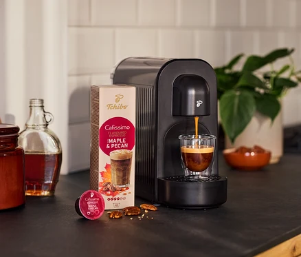 Čierny kávovar varí espresso do pohára. Vedľa balenie kapsúl s príchuťou javora a pekanových orechov, voľná kapsula, orechy, fľaša a rastlina.