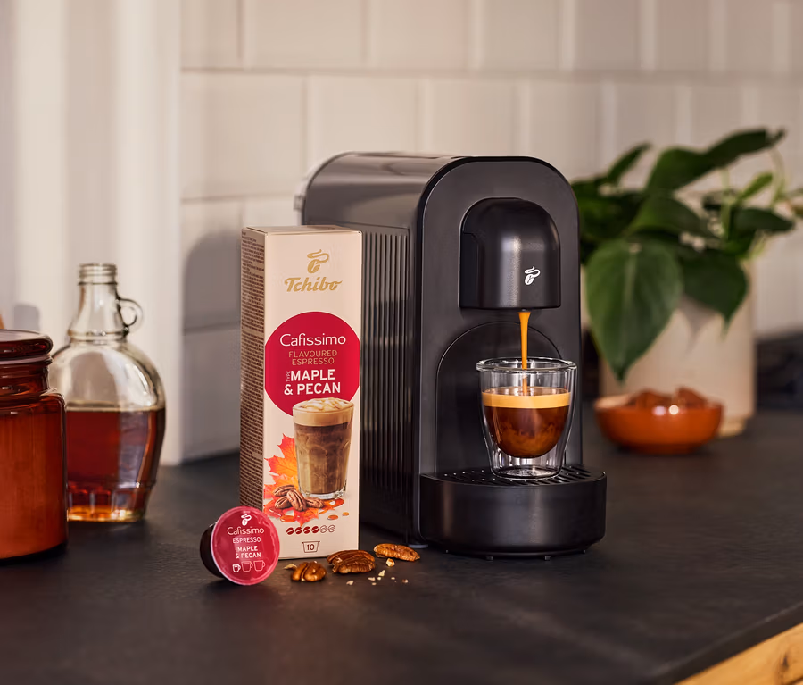 Čierny kávovar varí espresso do pohára. Vedľa balenie kapsúl s príchuťou javora a pekanových orechov, voľná kapsula, orechy, fľaša a rastlina.