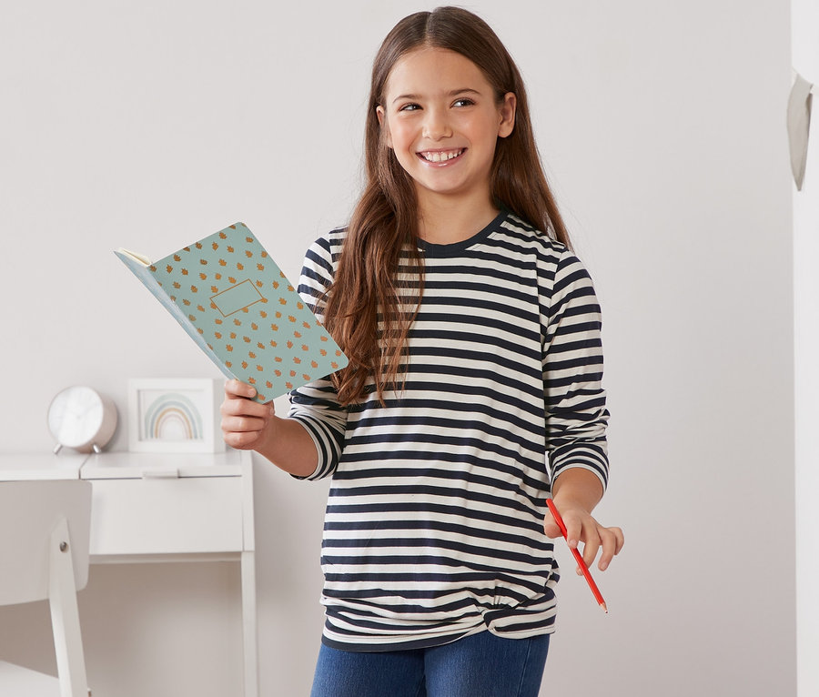 Dievča drží otvorený zápisník a červenú ceruzku. Má na sebe modro-biele prúžkované tričko s dlhým rukávom a zauzleným detailom.