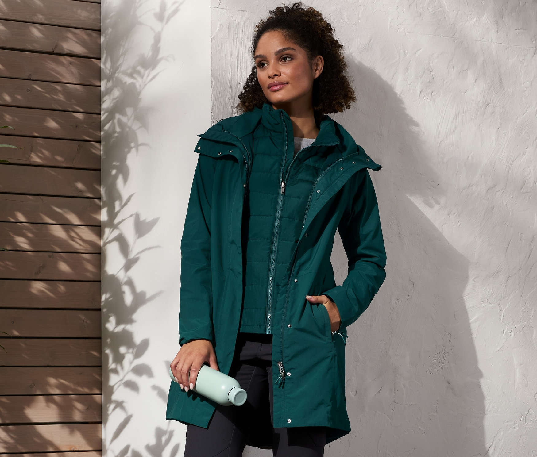 Žena stojí a má na sebe zelený kabát do dažďa s vestou, čierne nohavice a hnedé topánky. V ruke drží fľašu s vodou a vedľa nej stojí batoh.