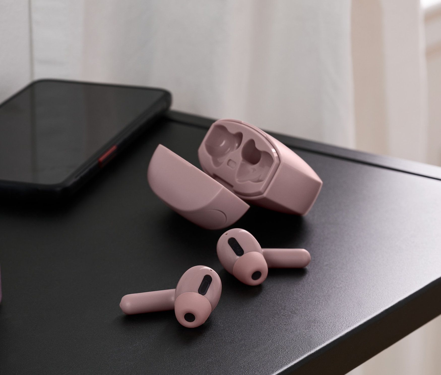 Na čiernom stolíku sú ružové slúchadlá do uší TWS, vedľa nich otvorené puzdro a mobilný telefón. V pozadí váza.