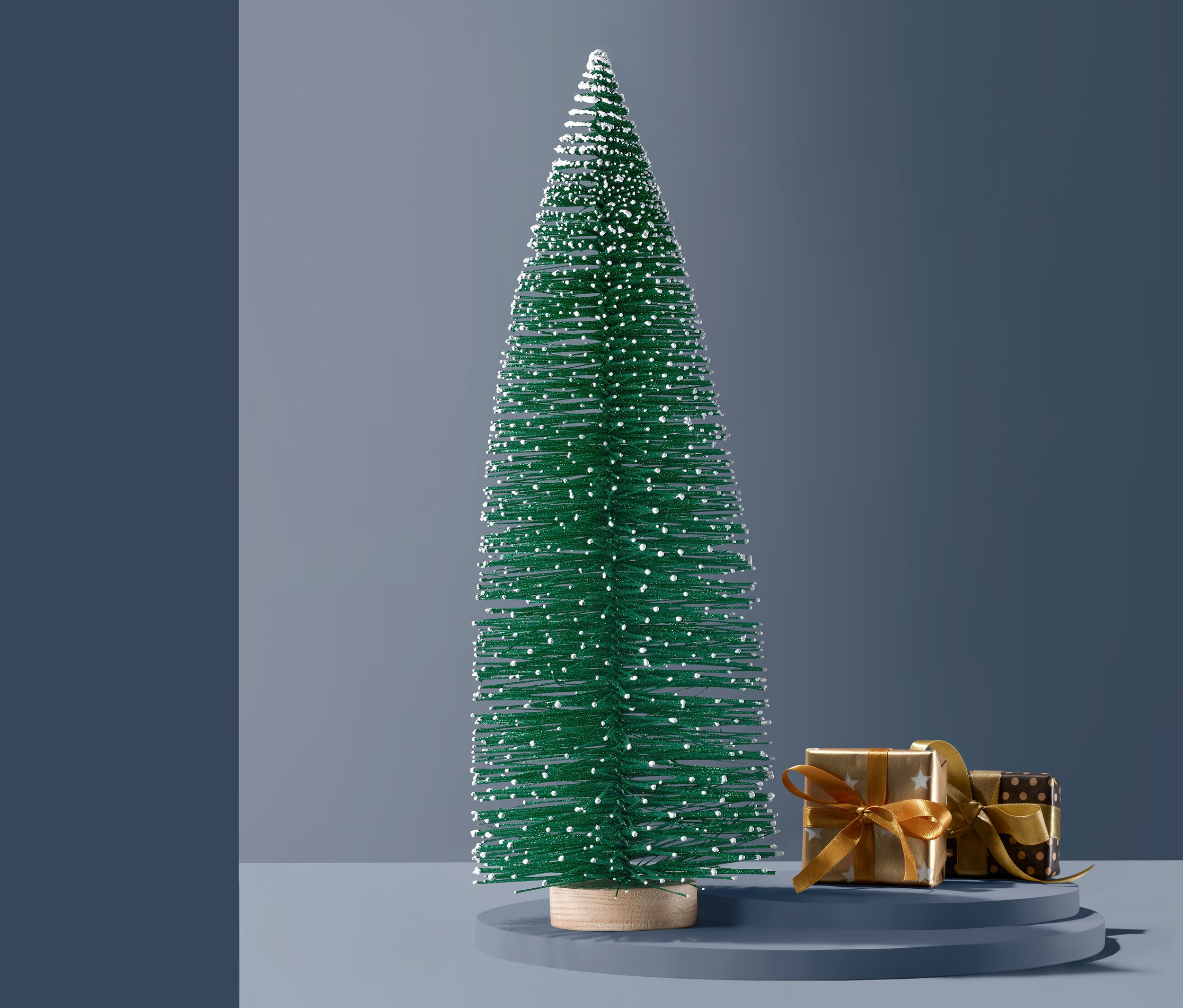 Zelený dekoračný stromček s bielymi škvrnami stojí vedľa dvoch zabalených darčekov so zlatými stuhami.