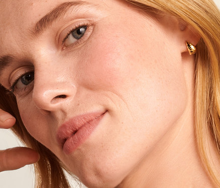 Blondínka s rukou pri hlave nosí retiazku Singapur zo zlata s rýdzosťou 585 a otvorené kreoly zo zlata s rýdzosťou 375, kónické, úprava diamantovaním.