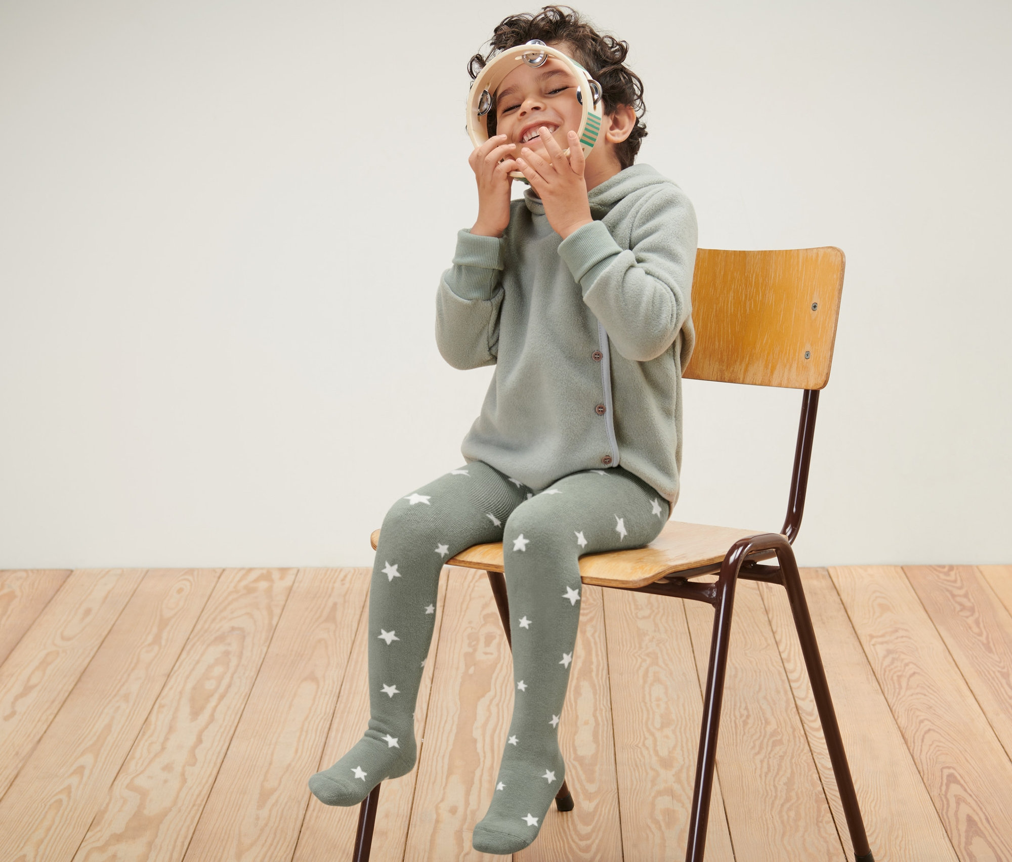 Chlapec sedí na stoličke a hrá sa s tamburínou. Má na sebe modré a zelené detské termo pančuchy.