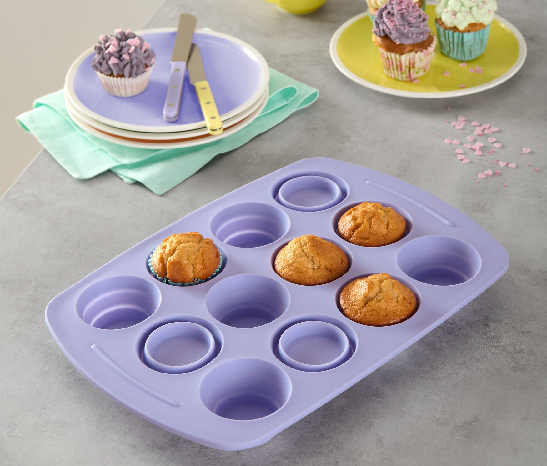 Fialová skladacia silikónová forma na muffiny s tromi muffiny. V pozadí sú taniere s muffiny, žltý hrnček a posýpka.
