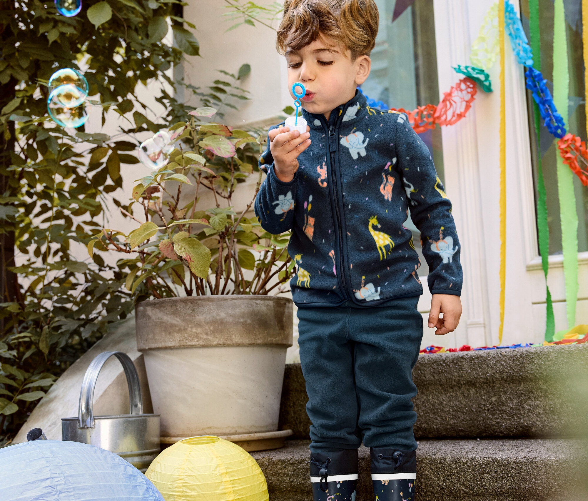 Chlapec fúka bubliny v detskej flísovej mikine, flísových nohaviciach a gumákoch.