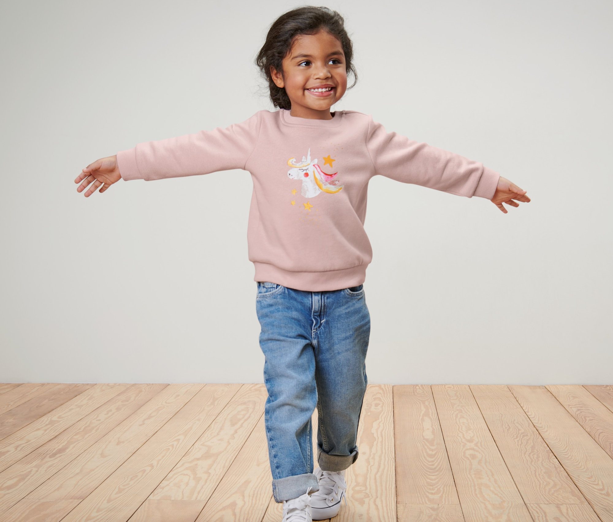 Dievča s rozpaženými rukami má na sebe detskú mikinu s jednorožcom, modré džínsy a tenisky.