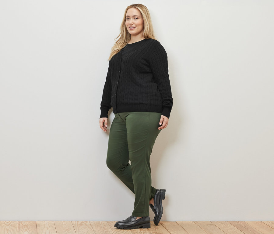 Žena pózuje v čiernom kardigáne z jemnej pleteniny s vrkočovým vzorom a zelených bengalínových nohaviciach.