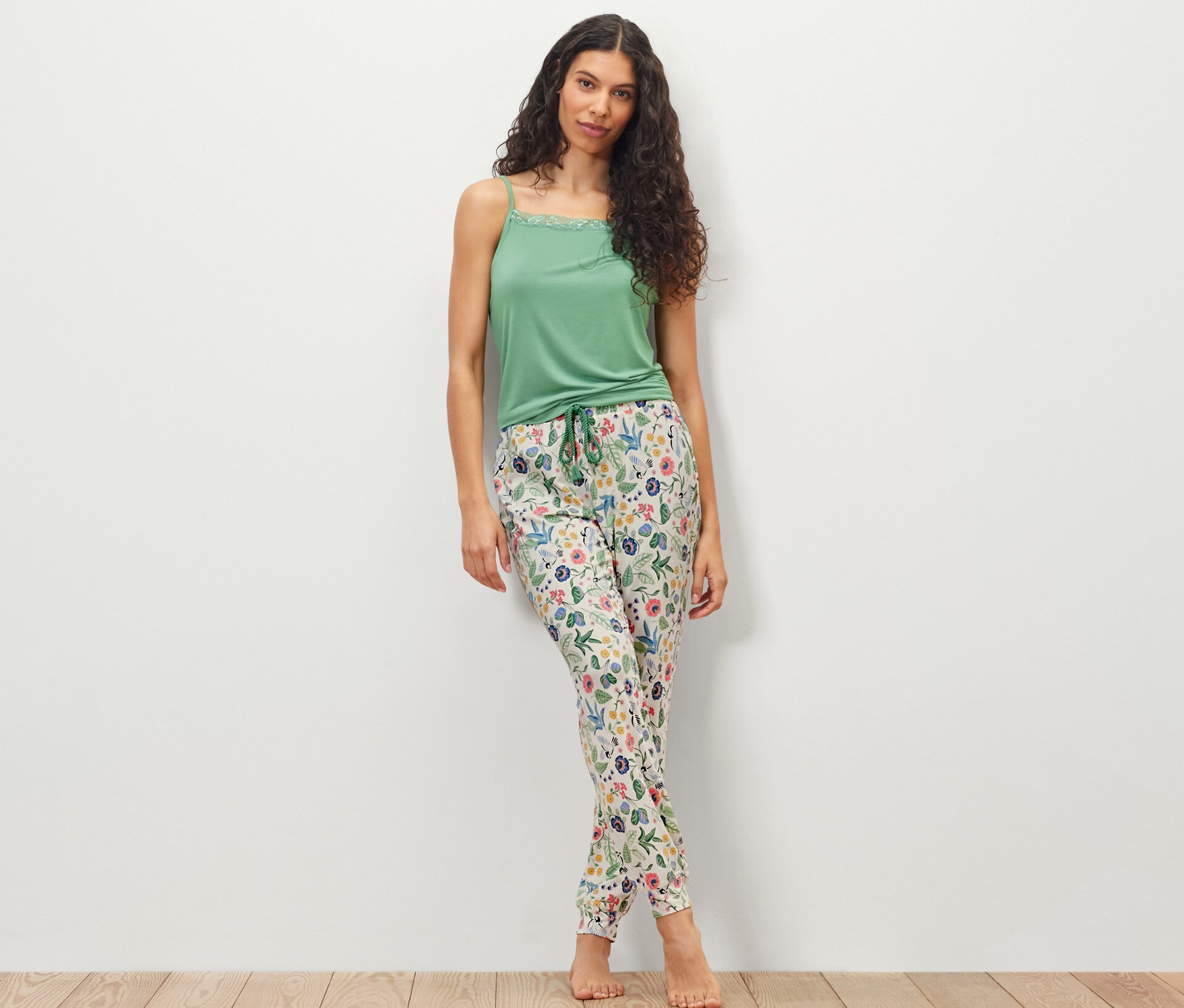 Žena stojaca pred bielou stenou má na sebe zelený top a pyžamové nohavice.