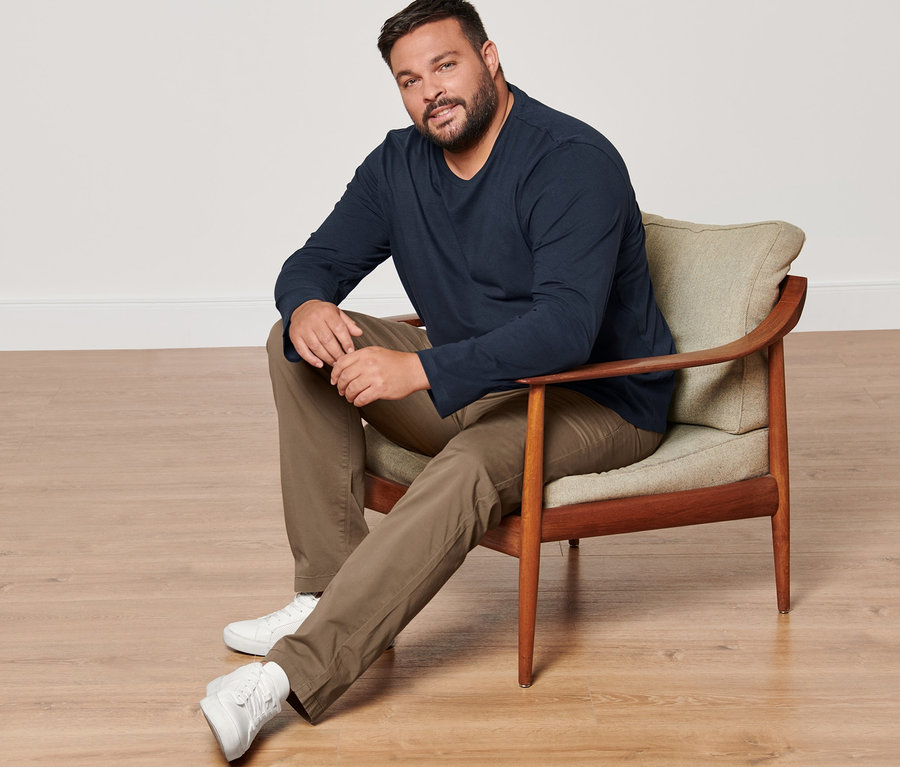 Muž sedí v kresle a má na sebe dve tričká s dlhým rukávom, pieskové nohavice chino a tenisky.