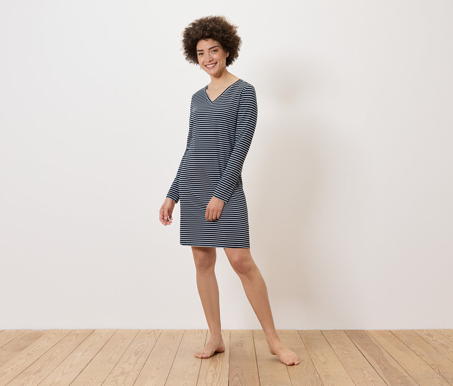 Žena stojí naboso na drevenej podlahe a má na sebe pruhované šaty s dlhým rukávom.