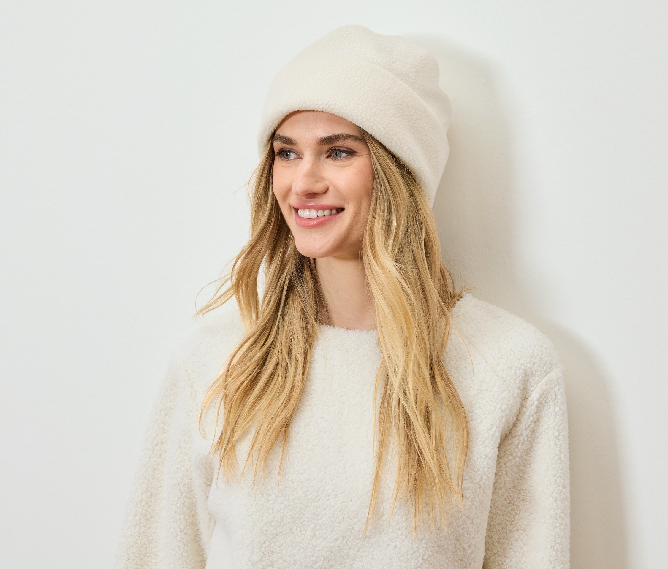 Usmievavá žena s blond vlasmi má na sebe krémovú čiapku a sveter.
