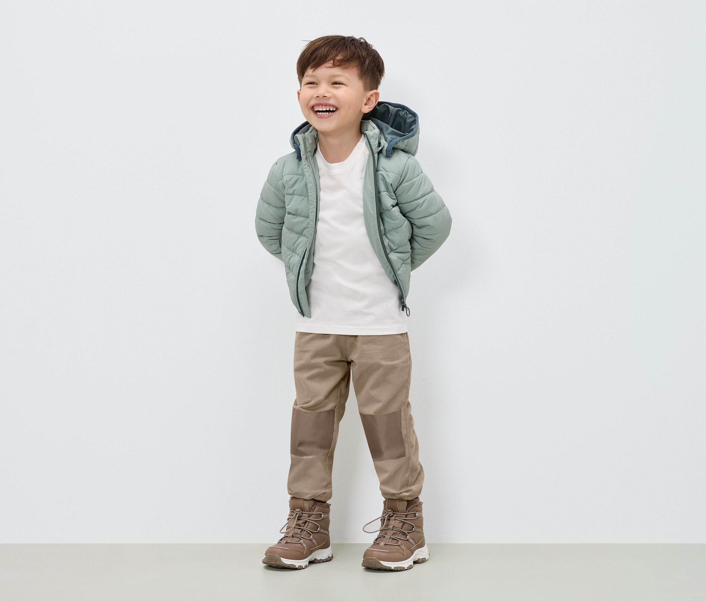 Usmievajúci sa chlapec stojí v svetlozelenej páperovej bunde, béžových nohaviciach a hnedých topánkach.