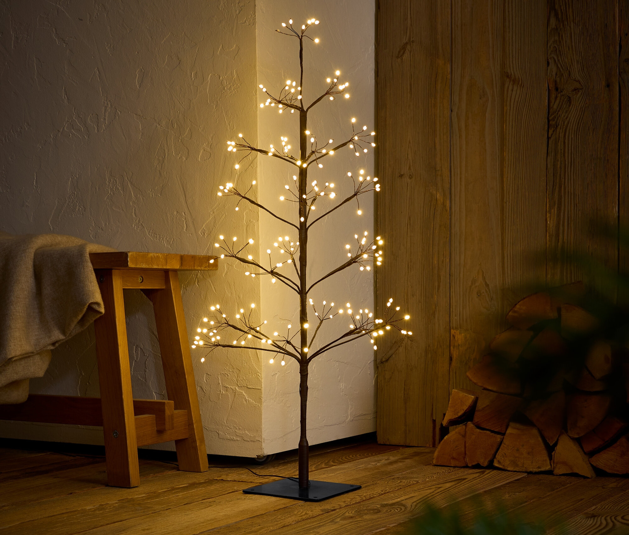 Dekoratívny strom s teplými bielymi svetlami stojí na drevenej podlahe vedľa lavice.