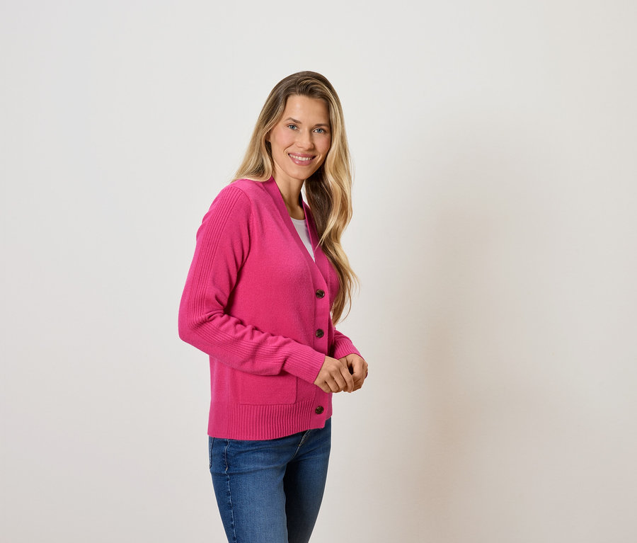 Žena s blond vlasmi pózuje v ružovom svetri a modrých džínsoch.